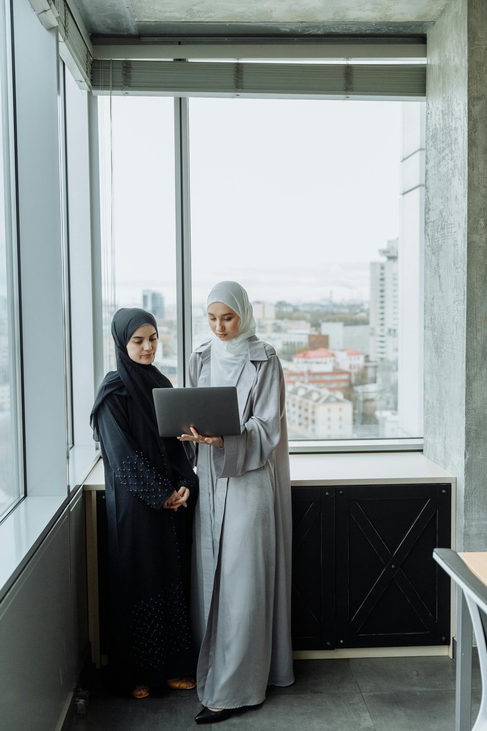 Two women in hijabs discussing work while standing by a window in a modern office setting.