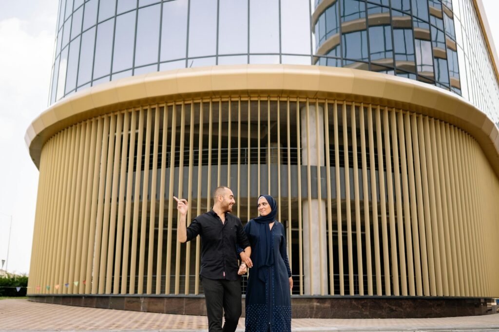 A couple walks hand-in-hand in front of a modern glass building, showcasing urban fashion.